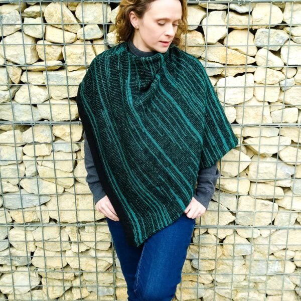 Model wearing teal and black knitted striped poncho, stood against a cream wall.