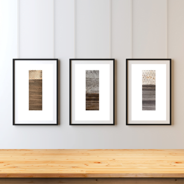 Su France Designs Earth prigment giclee trio - Three framed abstract artworks hang on a white wall above a light wooden floor. Each frame contains a vertical rectangular image divided into two sections. The top sections feature geometric or dotted patterns in light colors, while the bottom sections show darker, textured horizontal stripes resembling wood grain. The frames are black and evenly spaced, creating a cohesive triptych display.