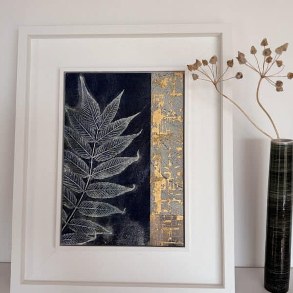 A leaf print in a white frame on a white shelf next to a vase of flowers