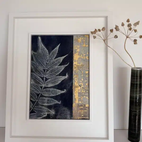 A leaf print in a white frame on a white shelf next to a vase of flowers