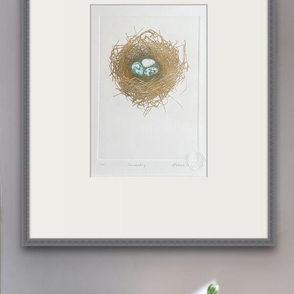 A framed solar plate print, by Su France Designs, hangs on a pale wall above stacked books. The print depicts a delicate bird's nest made of thin twigs, containing three small speckled blue eggs. The nest is rendered in golden tones against a white background. The print is matted in white and enclosed in a simple gray frame. Below the frame, several books are stacked on a surface, with a small plant visible on top of one book.