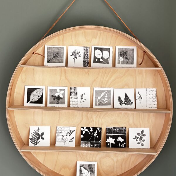 A circular wooden hanging shelf with three tiers displays a collection of small black and white botanical prints. The shelf is light wood, suspended by a leather cord against a gray-green wall. The prints are monoprints by Su France and feature various plant motifs including leaves, flowers, and ferns arranged neatly on each shelf level.