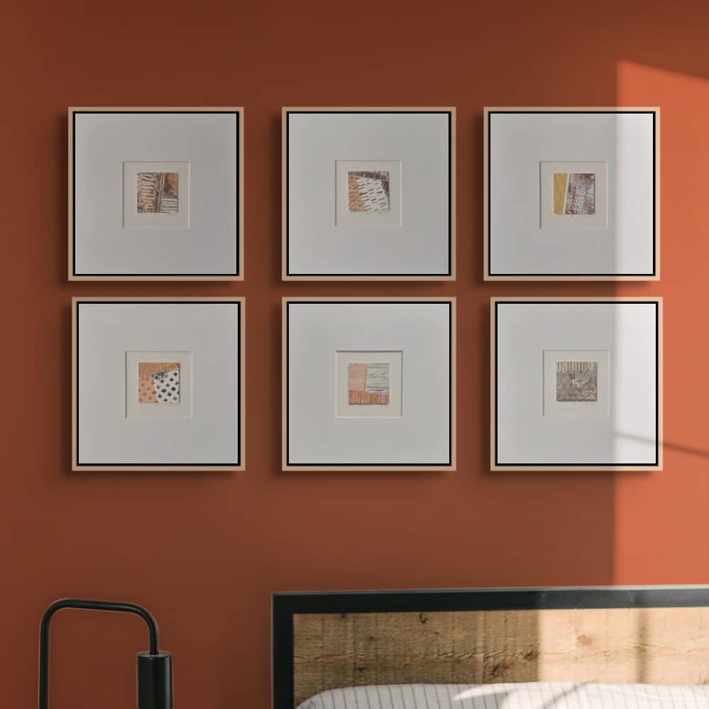 Six small collagraph prints in pale wooden frames with wide white mounts, hung in a grid of two rows of three on a terracotta wall above a bed. Each tiny print is a patchwork of stripes, dashes, lattice and dotted textures in burnt orange, peach, black, cream and ochre, looking like marks from fields.