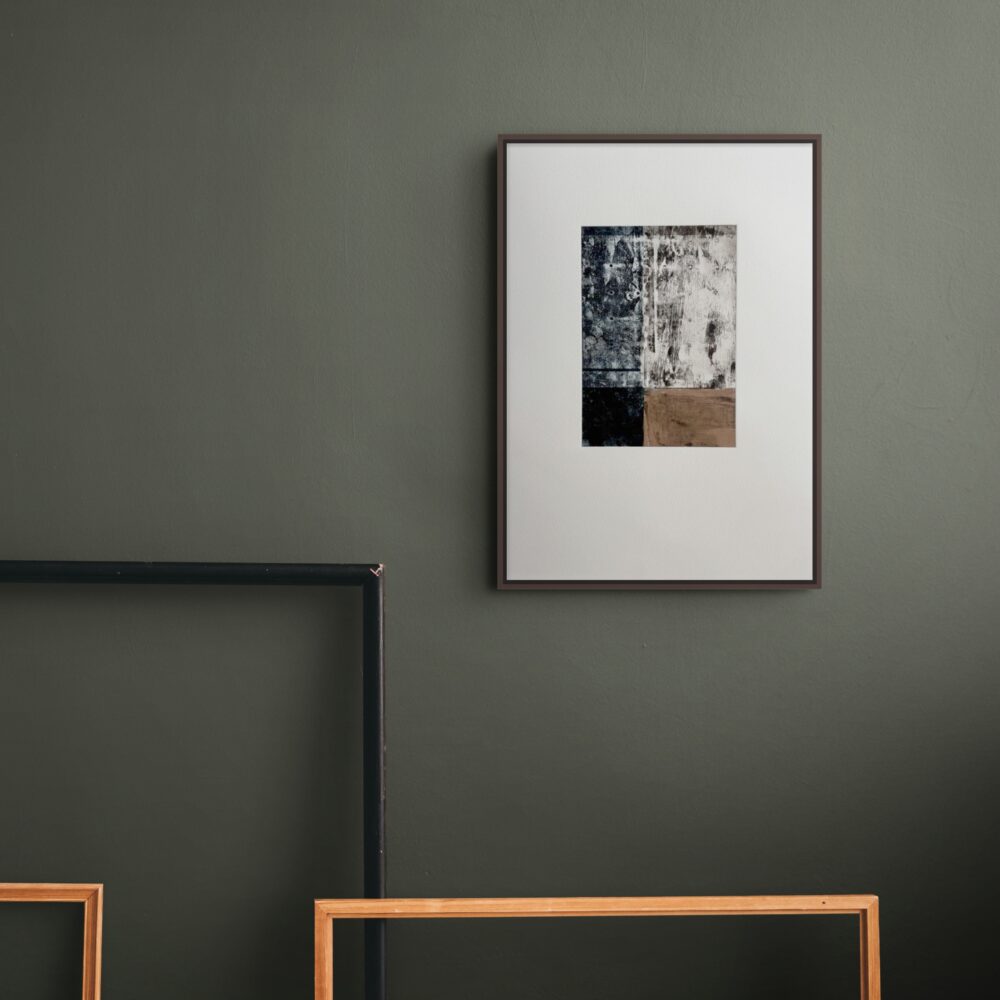 A framed abstract print in a dark brown frame with a wide white mount, hung on a deep olive green wall. The print is made of blocks of texture: inky blue and black marks, a pale weathered area with dripping, scratchy lines, and a patch of warm brown. Empty wooden and black frames lean in front of the wall.