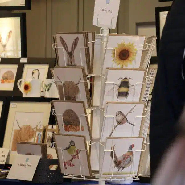 A stall selling wildlife artwork and cards with Customer browsing stalls in the background at Tissington Craft Fair in Derbyshire, Tissington Craft Fairs