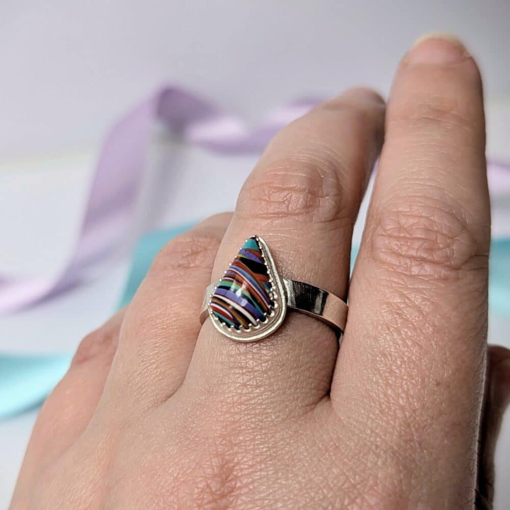 Image shows a hand wearing a chunky silver ring. the ring features a multicoloured teardrop shape surfite stone.