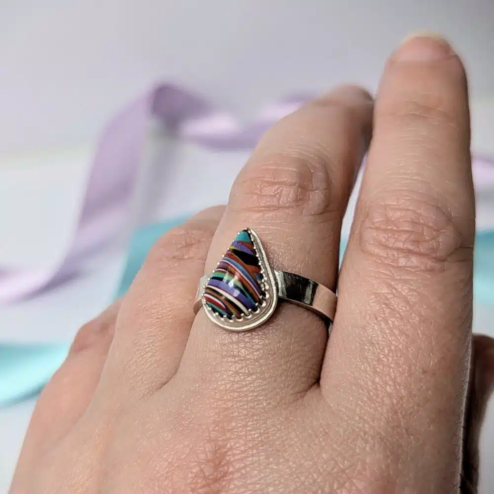 Image shows a hand wearing a chunky silver ring. the ring features a multicoloured teardrop shape surfite stone.