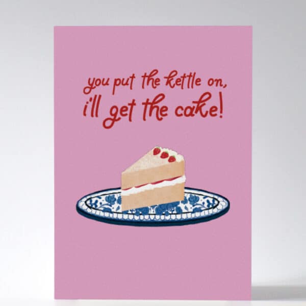 A product photo of a pink greeting card with a red custom typeface which reads, ‘you put the kettle on, I’ll get the cake!’ and the greeting card features a Multani pottery cake platter and a Victoria sponge cake slice.