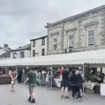 the image shows a large marquee in the high street, with artists selling their wares, with a large group of visitors. behnind th emarquee are the high street buildings
