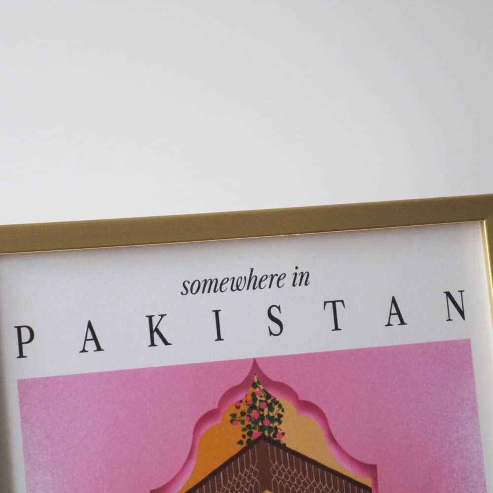 A close-up shot of a gold metallic frame with a colourful print with text 'somewhere in Pakistan' showing the top of an illustration of a pink Mughal arch and a haveli (mansion courtyard).
