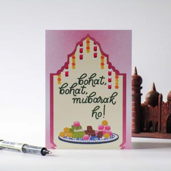 A product photo of a greeting card which reads 'bohat bohat mubarak ho!' which translates from Urdu or Hindi to many many congratulations. Behind the greeting card is a wooden sculpture prop of Badshahi Masjid, a well known masjid in Pakistan. There is also a pen as a prop beside the card.