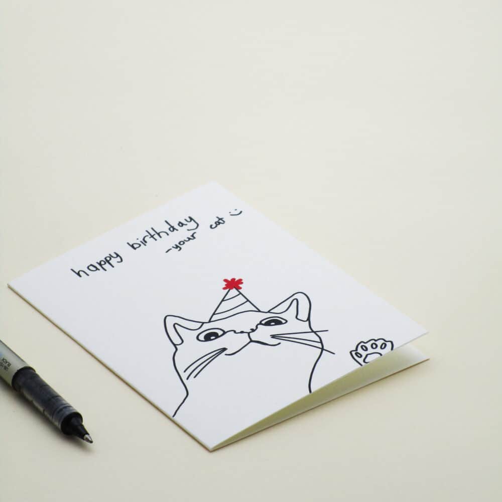 A product photo of a greeting card with custom handwritten words which read, ‘happy birthday - your cat :)’ and the greeting card features a simple line drawing of a cat smiling with its paw raised, wearing a party hat with a red sparkly decoration on the top.