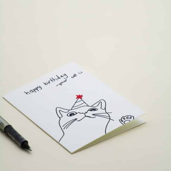 A product photo of a greeting card with custom handwritten words which read, ‘happy birthday - your cat :)’ and the greeting card features a simple line drawing of a cat smiling with its paw raised, wearing a party hat with a red sparkly decoration on the top.