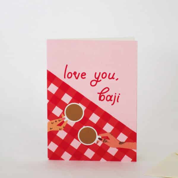 A product photo of a pink greeting card with a red custom typeface which reads, ‘love you, baji’ (where baji means sister in Urdu or Hindi) and the greeting card features a red and white gingham pattern as a tablecloth and two women holding their cups of tea in their hand. They are both wearing nail polish and mehndi / henna.