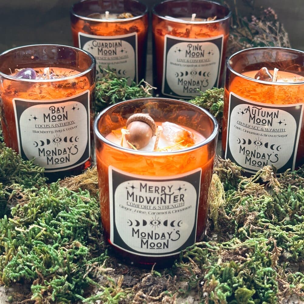 Image shows five candles in amber glass jars, dressed with an assortment of crystals and herbs on a bed of moss.