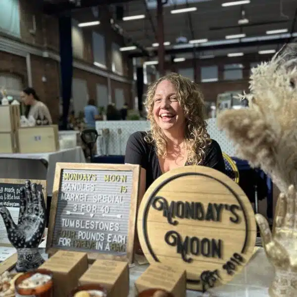 Image shows a woman with curly hair smiling behind a market stall.