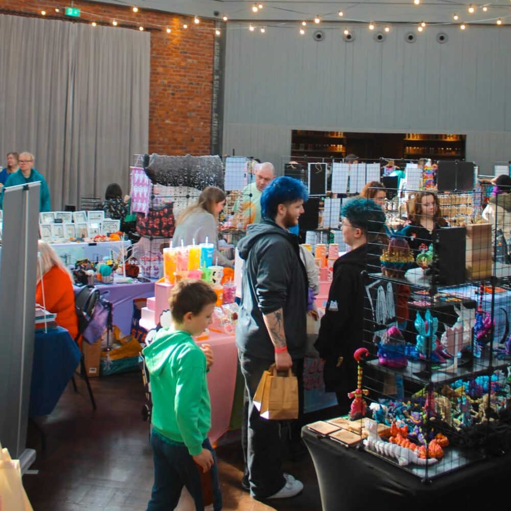 A room with brick walls and grey panelling filled with stalls and featuring some customers walking around. Infront of the camera is a little boy in a green hoodie and a person with bright blue hair