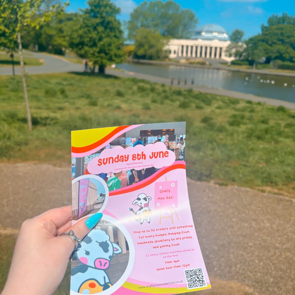 A hand holding a very colourful and bright pink flyer featuring some images and details about the market, the flyer is being held infront of the venue, Wylam Brewery with a lake and trees fatured in front of the venue, the sky is clear blue with some scattered clouds
