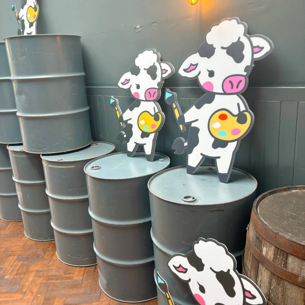 A grey hallway with grey metal barrels, which have cow cutouts on top of the barells. the cow is a cartoon style and is standing holding a paint brush and palette. The floor is rustic wooden.