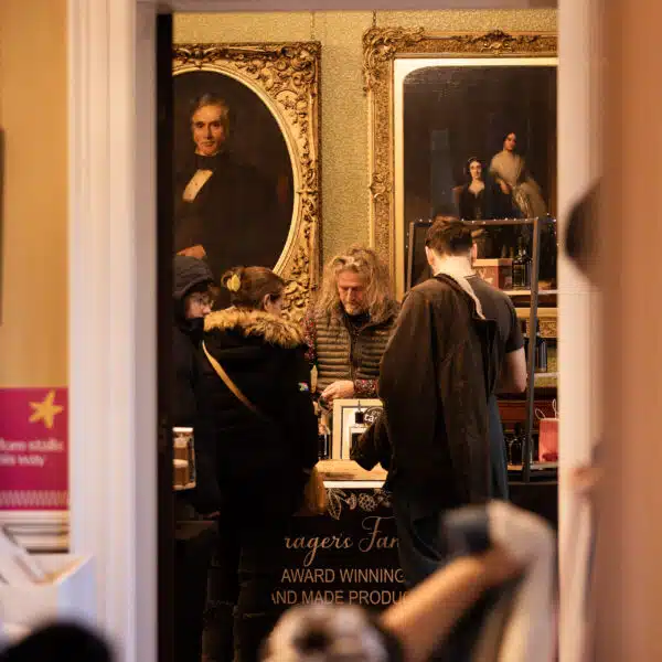 National Museums Liverpool - Pedddle - Image shows an open door, with a stallholder selling to customers. In the background, are two portraits from the Victorian era, and in the foreground there are more customers.