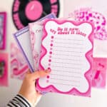 hand holding three colourful notepads designed by Snarki Studios. The front notepad features bold pink text that reads “Do it now, cry about it later” with a playful wavy border and bullet point lines. The other notepads show purple and pink designs with fun, quirky illustrations and phrases. In the background is a pink-themed wall with vinyl records and art prints, creating a vibrant, creative atmosphere.