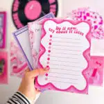 hand holding three colourful notepads designed by Snarki Studios. The front notepad features bold pink text that reads “Do it now, cry about it later” with a playful wavy border and bullet point lines. The other notepads show purple and pink designs with fun, quirky illustrations and phrases. In the background is a pink-themed wall with vinyl records and art prints, creating a vibrant, creative atmosphere.
