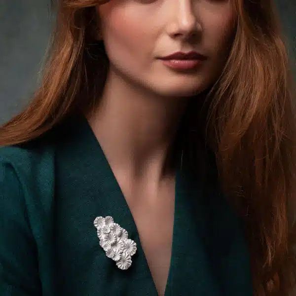A model in a green dress wears a beautiful silver brooch made of a cluster of silver flower like shapes sculpted using real poppy seedheads
