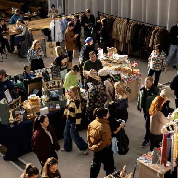 Campfield Market: A high-angle shot captures a busy indoor market hall filled with people browsing various vendor stalls. Stalls are arranged throughout the open space, featuring items such as honey and jars of jam, baked goods, vintage clothing on racks, and colorful scarves. To the left, several people are seated at long wooden tables in a dining area. The space is bright, illuminated by soft light coming from the side.