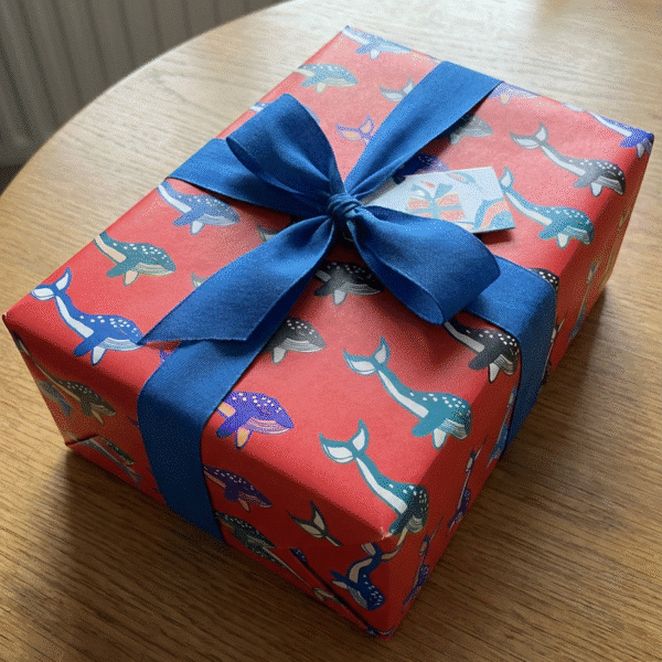 Juicy Prints - A gift on a wooden table wrapped in red wrapping paper with whale illustrations printed on it and big blue bow.