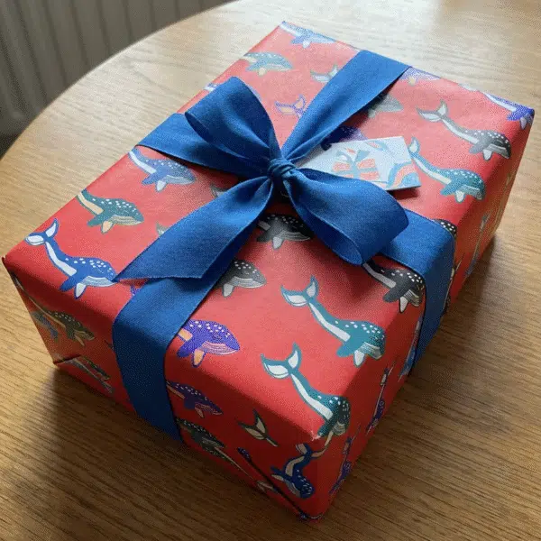A gift on a wooden table wrapped in red wrapping paper with whale illustrations printed on it and big blue bow.