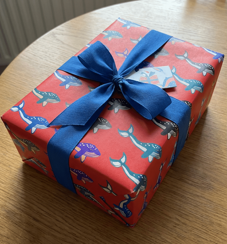 Juicy Prints - A gift on a wooden table wrapped in red wrapping paper with whale illustrations printed on it and big blue bow.