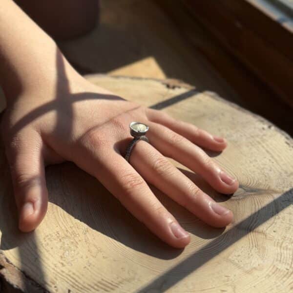 A molten cup ring from the Alchemy range. It is oxidised with a white effect inside the cup part of the ring. Worn by model on hand resting on wood by a window.