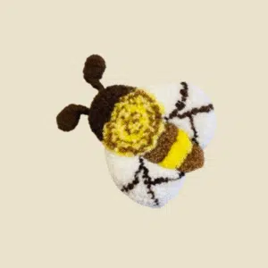 Handmade punch needle bee with white wings, yellow stripes, and brown details on a neutral background.Handmade textile bee, a perfect addition to a collection of decorative wall crafts.