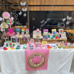 A brightly lit indoor craft market stall for "Rose Weaver Tales". The table is covered in a white tablecloth with a pink banner featuring a rose logo hanging in the center. The display is filled with colorful, textured punch needle and tufted textile art items, including large moths and bee wall hangings on the left, small potted flower decorations, square framed fiber art pieces on a wooden riser, and dozens of colorful round coasters, jewelery trays and keychains spread across the tabletop. Small pink price tags are placed next to the items.