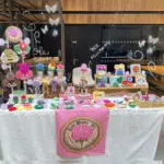 A brightly lit indoor craft market stall for "Rose Weaver Tales". The table is covered in a white tablecloth with a pink banner featuring a rose logo hanging in the center. The display is filled with colorful, textured punch needle and tufted textile art items, including large moths and bee wall hangings on the left, small potted flower decorations, square framed fiber art pieces on a wooden riser, and dozens of colorful round coasters, jewelery trays and keychains spread across the tabletop. Small pink price tags are placed next to the items.