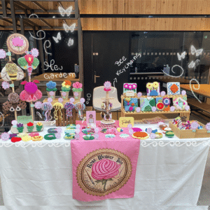 A brightly lit indoor craft market stall for "Rose Weaver Tales". The table is covered in a white tablecloth with a pink banner featuring a rose logo hanging in the center. The display is filled with colorful, textured punch needle and tufted textile art items, including large moths and bee wall hangings on the left, small potted flower decorations, square framed fiber art pieces on a wooden riser, and dozens of colorful round coasters, jewelery trays and keychains spread across the tabletop. Small pink price tags are placed next to the items.