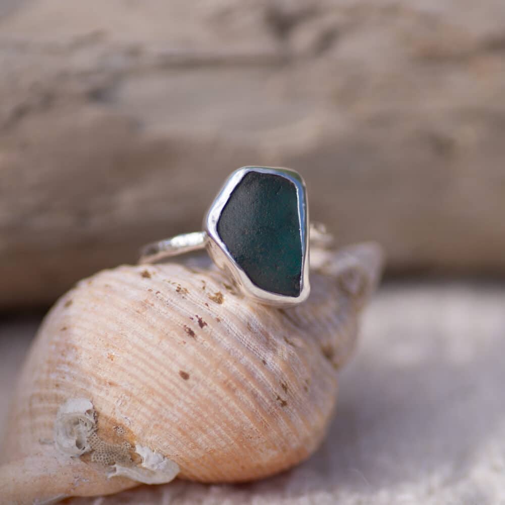 Aqua sea glass sterling silver ring with a textured band, sitting on the cone of a sea shell with driftwood in the background