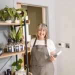 Innerlather: a pciture of Nicci Trudgeon holding a book of recipes standing by a wooden shelf of her products