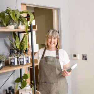 Innerlather: a pciture of Nicci Trudgeon holding a book of recipes standing by a wooden shelf of her products