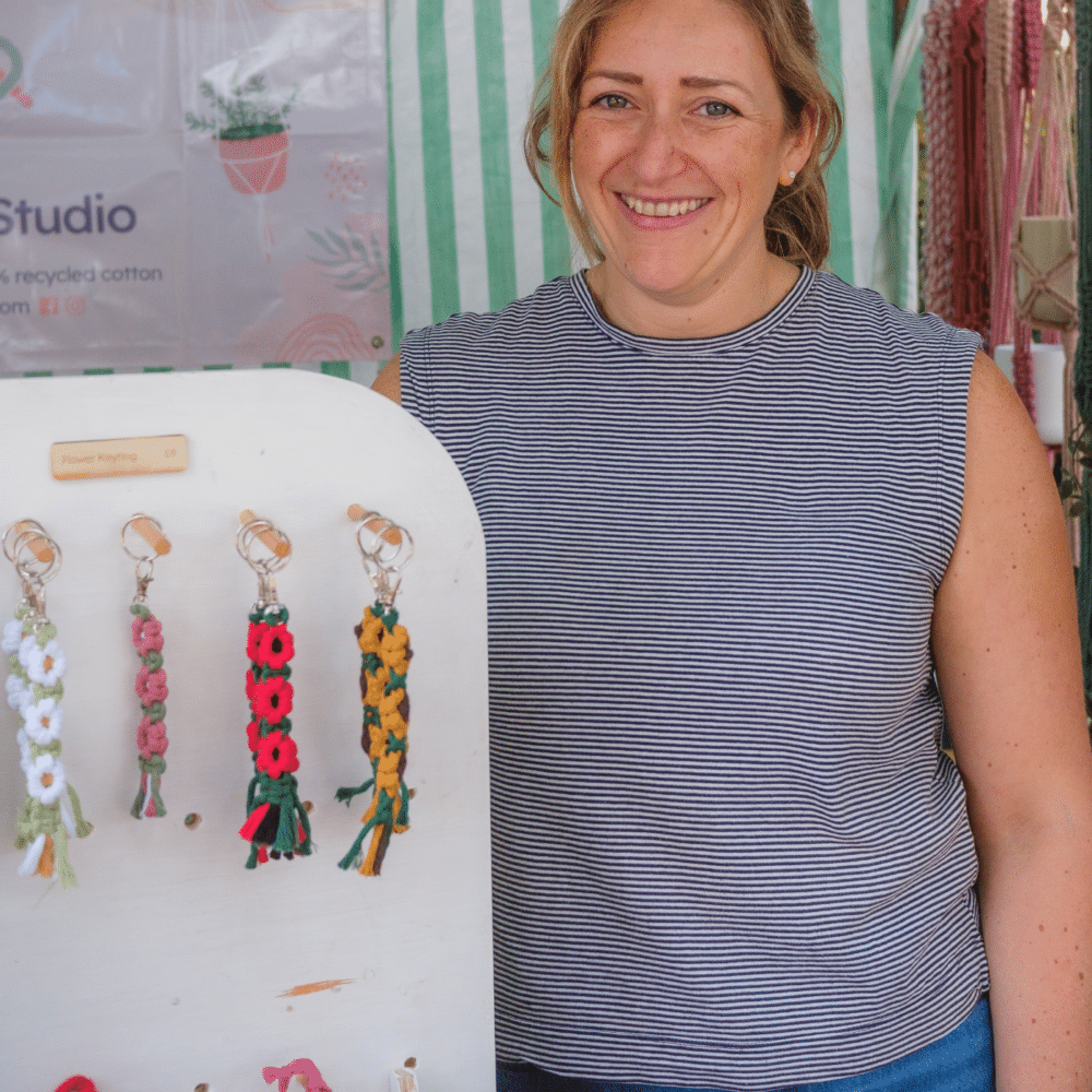 Sam, the maker from Sambrini Studio at a market displaying handmade flower keyrings and colourful macramé plant hangers.